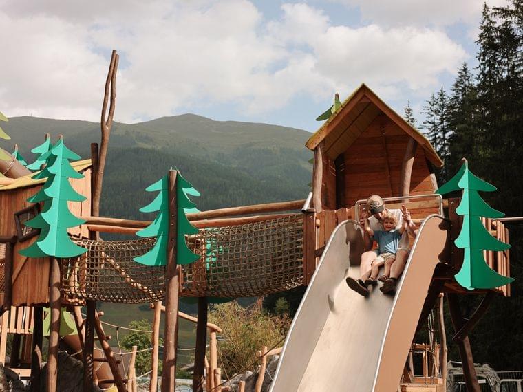 Turm mit Edelstahlbreitrutsche und Rutschstange am Habachklause Spielplatz Vater rutscht mit seinem Kind über eine breite Edelstahlrutsche am Spielplatz